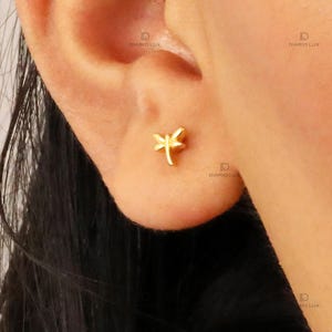 May include: A small gold-toned stud earring shaped like a dragonfly. The earring features a central body and two pairs of wings extending outward, positioned directly on the earlobe. The design is simple, metallic, and polished in appearance.
