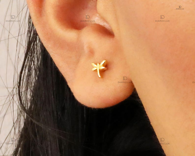 May include: A small gold-toned stud earring shaped like a dragonfly. The earring features a central body and two pairs of wings extending outward, positioned directly on the earlobe. The design is simple, metallic, and polished in appearance.