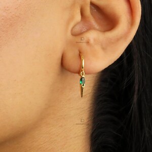 May include: A gold hoop earring featuring a small, teardrop-shaped green gemstone pendant. The earring is thin and delicate, with a polished metallic finish. The text DIARIO LUX is visible in small, faint lettering near the earring.