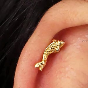 May include: Gold fish shaped cartilage earring. The earring is a small, detailed fish with a textured surface.