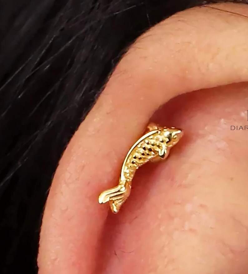 May include: Gold fish shaped cartilage earring. The earring is a small, detailed fish with a textured surface.