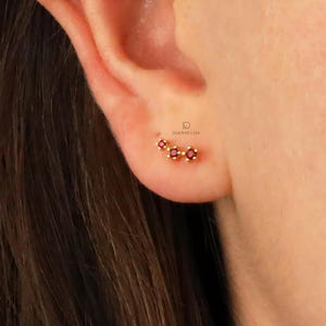 May include: A close-up of a person's ear with a small gold earring featuring three red gemstones. The earring is shaped like a crescent moon.