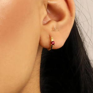 Może przedstawiać: Złote kolczyki koła z małym czerwonym kamieniem szlachetnym. Kolczyki są noszone w uchu osoby.