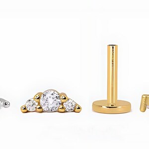 Op de afbeelding: Een set lichaamsieraden bestaande uit een zilverkleurige knop met platte achterkant en een top met drie stenen, een goudkleurige top met drie stenen, een goudkleurige staaf met platte achterkant en een goudkleurige top met schroefdraad.
