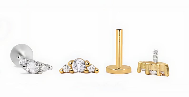 Op de afbeelding: Een set lichaamsieraden bestaande uit een zilverkleurige knop met platte achterkant en een top met drie stenen, een goudkleurige top met drie stenen, een goudkleurige staaf met platte achterkant en een goudkleurige top met schroefdraad.