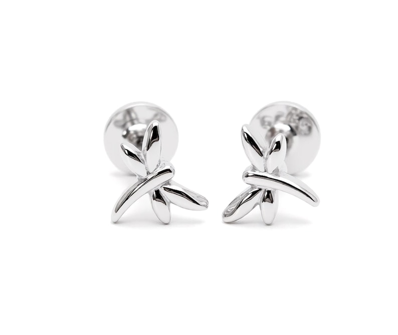 May include: A pair of silver-toned stud earrings shaped like dragonflies. Each earring features a polished, reflective surface with smooth, curved wings and a slender body design. The studs include circular backings visible behind the dragonfly motif.