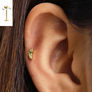May include: A gold-tone flat back stud earring featuring three small, round, light green stones arranged in a curved cluster. The earring is shown inserted into the cartilage of an ear, with a separate diagram showing the post and screw mechanism.