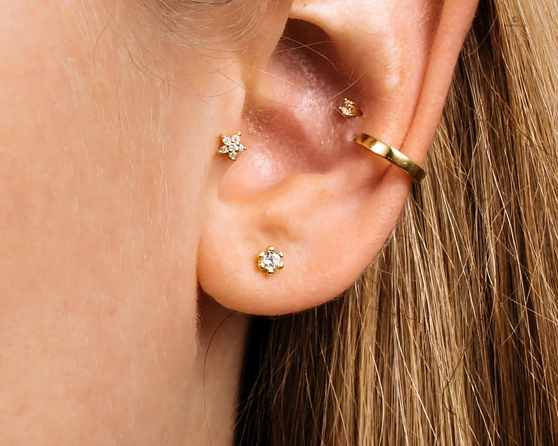 May include: A close-up of a person's ear with three gold earrings. The earrings are a small round stud, a larger flower-shaped stud, and a gold hoop.