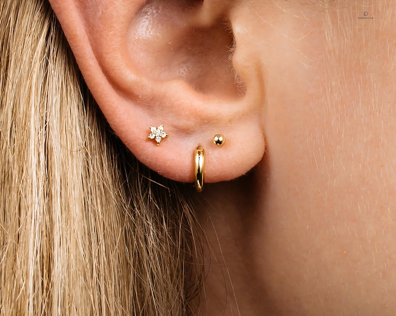 May include: A close-up of a person's ear with three gold earrings. The earrings are a small flower with clear stones, a small gold ball, and a gold hoop.