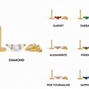 Op de afbeelding: Een gouden labret-piercing met platte achterkant en een gebogen topje met drie stenen. De set toont verschillende edelsteenopties met de labels Diamond, Garnet, Emerald, Alexandrite, Peridot, Pink Tourmaline en Sapphire, elk met een bijpassende stift.