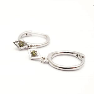 Pode incluir: Brincos de argola de prata com um pingente em forma de diamante com duas pedras preciosas verdes.