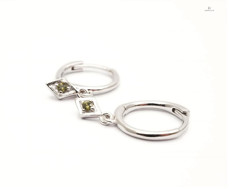 Pode incluir: Brincos de argola de prata com um pingente em forma de diamante com duas pedras preciosas verdes.