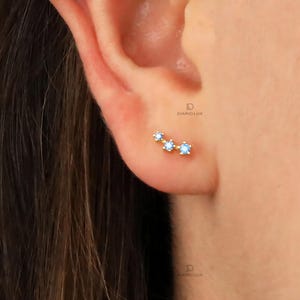 May include: A close-up of a person's ear with a small gold earring featuring three light blue gemstones. The earring is shaped like a crescent moon.