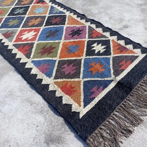 May include: A black and white geometric patterned rug with colourful accents. The rug has a fringe border and is made of wool.