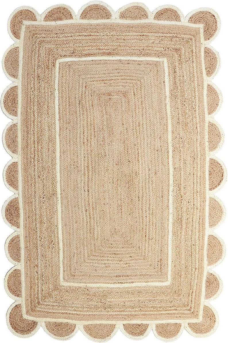 May include: A rectangular jute rug with a scalloped edge. The rug has a natural tan colour with a cream-coloured border and a rectangular design in the centre. The rug's dimensions are not visible.