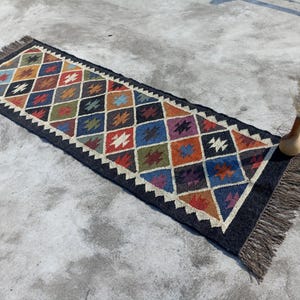 May include: A long, rectangular rug with a black background and a colourful geometric pattern. The rug has a fringe border and is made of natural fibres.