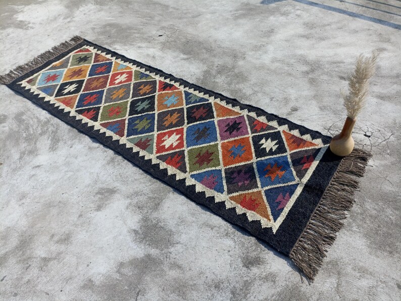 May include: A long, rectangular rug with a black background and a colourful geometric pattern. The rug has a fringe border and is made of natural fibres.