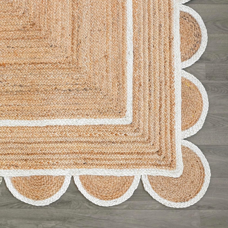 May include: A natural jute rug with a scalloped edge. The rug features a square centre with a white border and a series of semi-circular shapes along the edge. The rug's texture is visible, showcasing the woven fibres.