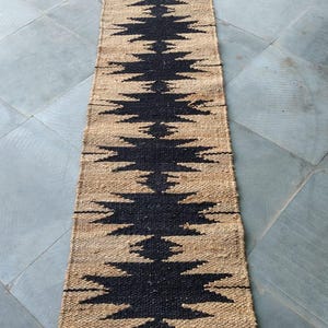 Op de afbeelding: Een lang, smal jutekleed met een geometrisch patroon in zwart en natuurlijke kleur. Het patroon is een reeks herhalende driehoeken.