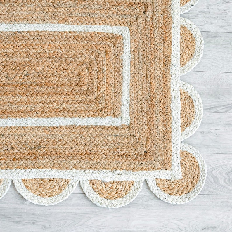 May include: A natural fibre rug with a braided texture and a scalloped edge. The rug features a rectangular centre with a white border, and the outer edge has a series of semi-circular shapes. The rug is made of a light brown material.