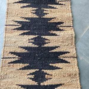 Op de afbeelding: Een vloerkleed van natuurlijke vezels met een zwart en bruin geometrisch patroon. Het vloerkleed is geweven met een textuur, een licht ruw oppervlak.