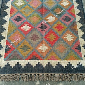 May include: A colorful, handwoven rug with a geometric pattern in shades of red, blue, brown, and white. The rug has a black border and fringe on the ends. The text "BhojArtCrafts" is visible on the rug.