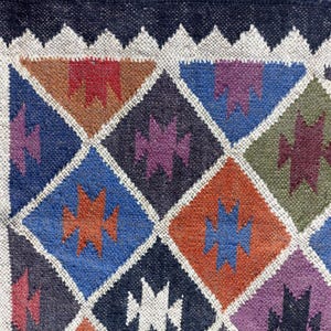 May include: A close-up of a colourful woven rug with a geometric pattern of diamonds and triangles. The rug is made of wool and has a dark blue border.