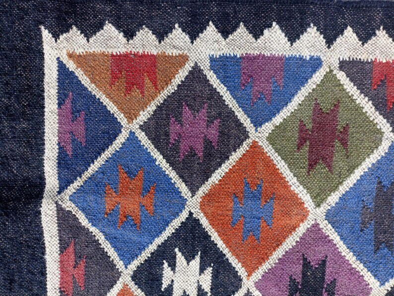 May include: A close-up of a colourful woven rug with a geometric pattern of diamonds and triangles. The rug is made of wool and has a dark blue border.