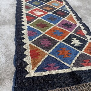 May include: A long, narrow rug with a geometric pattern in shades of blue, red, orange, green, and brown. The rug has a black background and a fringe border.