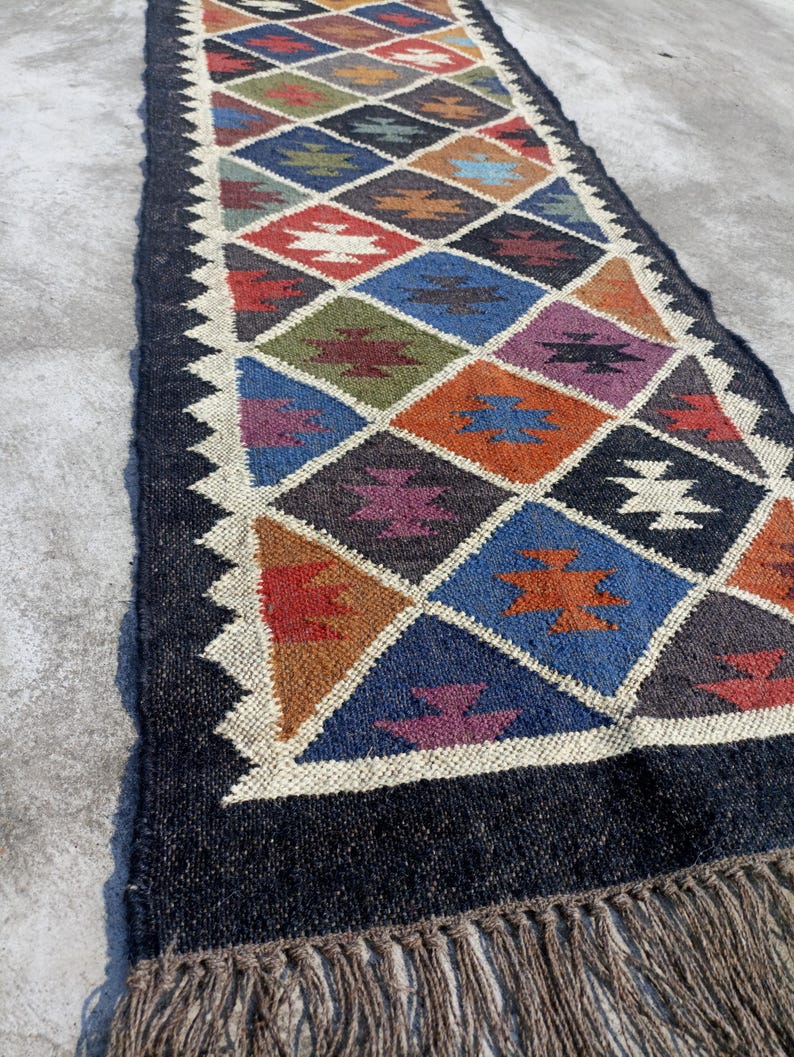 May include: A long, narrow rug with a geometric pattern in shades of blue, red, orange, green, and brown. The rug has a black background and a fringe border.