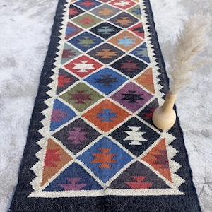 May include: A colourful, geometric patterned rug with a black background and fringe on the ends. The rug is made of wool and has a traditional design.