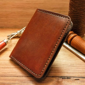 May include: A brown leather wallet with stitched edges. The wallet is closed and lying on a wooden surface.