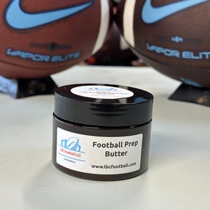 Könnte beinhalten: Ein kleines Glas Football Prep Butter, ein Produkt zur Vorbereitung von Fußbällen. Das Glas ist schwarz mit einem weißen Etikett, auf dem "Football Prep Butter" und "www.lbcfootball.com" steht.
