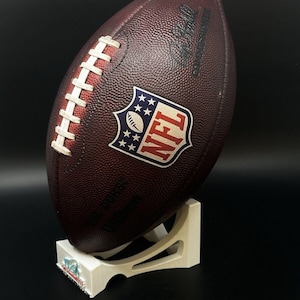 May include: A brown leather NFL football with white laces and the NFL logo, displayed on a white stand. The football is oval-shaped and has a textured surface. The stand has a team logo on the base.