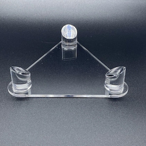 Könnte beinhalten: Transparenter Acryl-Präsentationsständer mit dreieckigem Design. Er verfügt über drei zylindrische Stützen und eine horizontale Basis. Der Ständer dient dazu, Artikel zu präsentieren und bietet eine elegante und minimalistische Präsentation.