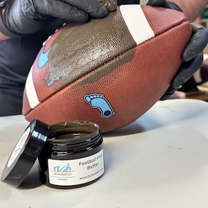 May include: A brown jar with a black lid labeled "Football Prep Butter" and the website "www.lbcfootball.com". The jar is open and a gloved hand is applying the brown butter to a brown and white football.