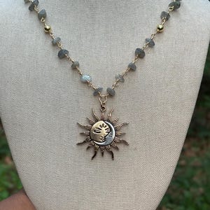 Può includere: Una collana color oro con un ciondolo a forma di sole e luna. Il ciondolo è decorato con un volto e raggi. La collana ha una catena di piccole perle di pietre preziose grigie e dettagli color oro. La collana è esposta su un manichino color neutro.