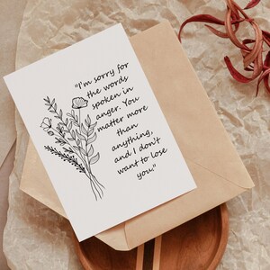 Puede incluir: Una tarjeta blanca con una ilustración floral y el texto "I'm sorry for the words spoken in anger. You matter more than anything, and I don't want to lose you." está colocada sobre un sobre marrón.