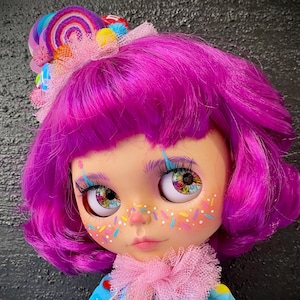 May include: A custom doll with vibrant purple hair and colorful eye details. The doll has a blue dress with candy accents and a pink ruffled collar. A candy-themed headpiece with a rainbow swirl and colorful accents completes the look.