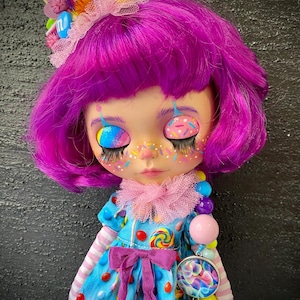 May include: A custom doll with purple hair and whimsical makeup, including rainbow-colored eyelids and freckles. The doll wears a blue dress adorned with candy and a pink bow. A candy-themed headband and necklace complete the look.