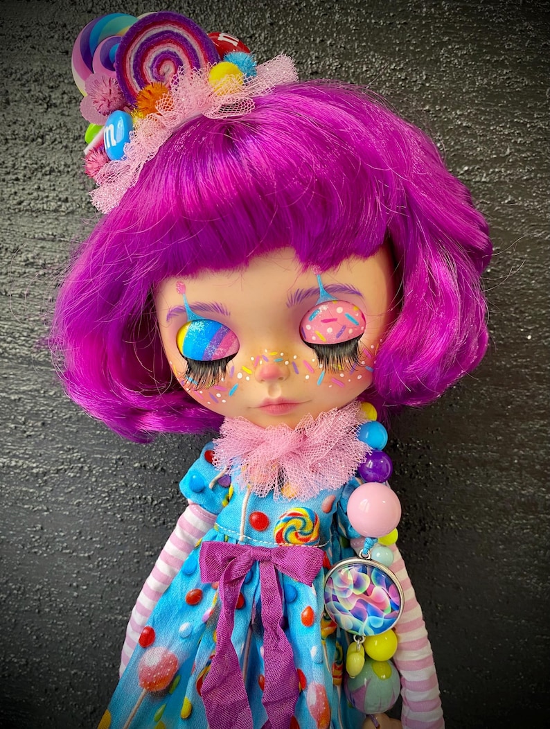 May include: A custom doll with purple hair and whimsical makeup, including rainbow-colored eyelids and freckles. The doll wears a blue dress adorned with candy and a pink bow. A candy-themed headband and necklace complete the look.