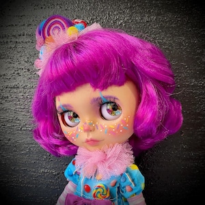 May include: A custom doll with vibrant purple hair and colorful eye details. The doll has a candy-themed outfit, including a blue dress with candy accents and a pink ruffled collar. A candy-themed headband completes the look.