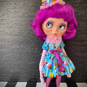 May include: A doll with bright purple hair and a candy-themed outfit. The doll wears a blue dress adorned with colorful candies and a matching candy-topped headpiece. Pink tights, striped sleeves, and blue shoes complete the whimsical look. The doll has large, expressive eyes.