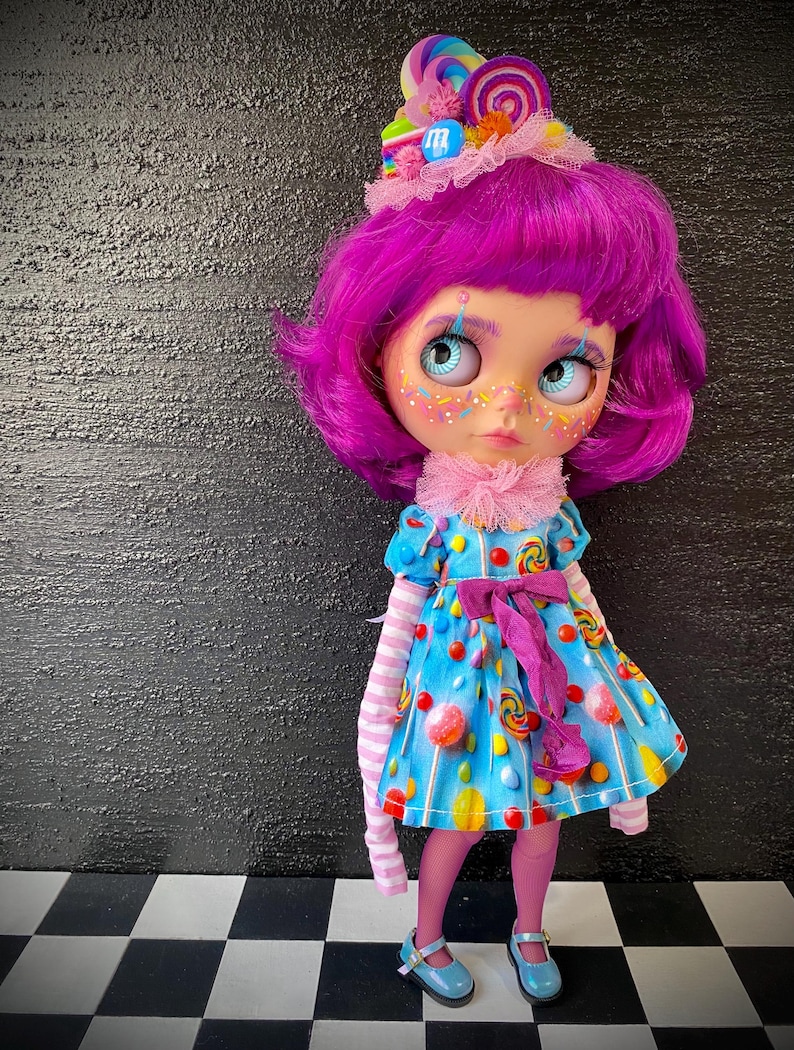 May include: A doll with bright purple hair and a candy-themed outfit. The doll wears a blue dress adorned with colorful candies and a matching candy-topped headpiece. Pink tights, striped sleeves, and blue shoes complete the whimsical look. The doll has large, expressive eyes.
