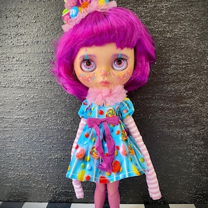 May include: A custom doll with purple hair and a candy-themed outfit. The doll wears a blue dress with candy prints, a pink bow, and striped sleeves. A candy-decorated headpiece and pink tights complete the look. The doll has painted freckles and large eyes.