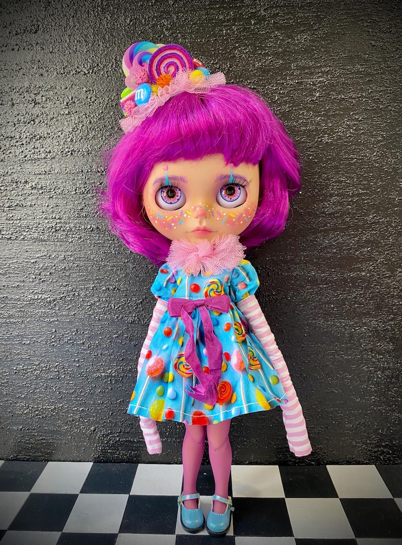 May include: A custom doll with purple hair and a candy-themed outfit. The doll wears a blue dress with candy prints, a pink bow, and striped sleeves. A candy-decorated headpiece and pink tights complete the look. The doll has painted freckles and large eyes.
