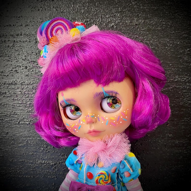 May include: A custom doll with vibrant purple hair and colorful eye details. The doll has a blue dress with candy accents and a pink ruffled collar. A candy-themed headpiece with a rainbow swirl and colorful accents completes the look.