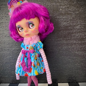 May include: A custom doll with vibrant pink hair and a matching lollipop hair accessory. The doll wears a blue dress adorned with candy graphics, a pink bow, and striped sleeves. She has blue shoes and pink tights.