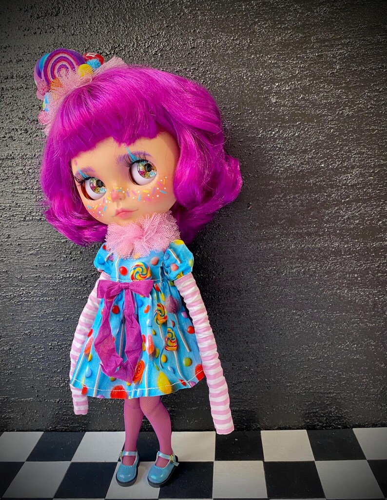 May include: A custom doll with vibrant pink hair and a matching lollipop hair accessory. The doll wears a blue dress adorned with candy graphics, a pink bow, and striped sleeves. She has blue shoes and pink tights.