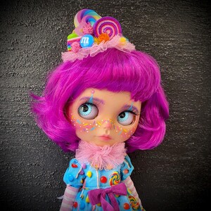 May include: A doll with bright purple hair and blue eyes, adorned with a candy-themed headdress. The doll wears a blue dress decorated with candy and a pink ruffled collar. The face is decorated with colorful sprinkles.